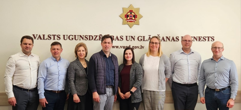 Pieci vīrieši un trīs sievietes vienā rindā stāv un skatās fotogrāfa virzienā. Fonā uz sienas redzama VUGD emblēma un uzraksts bordo krāsā: Valsts ugunsdzēsības un glābšanas dienests
