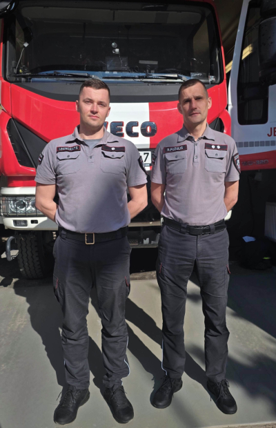 Divi pelēkos formastērpos tērpti Valsts ugunsdzēsības un glābšanas dienesta nodarbinātie stāv viens otram blakus un skatās fotogrāfa virzienā. Aiz viņiem redzama sarkani balta ugunsdzēsēju glābēju automašīnas priekšpuse