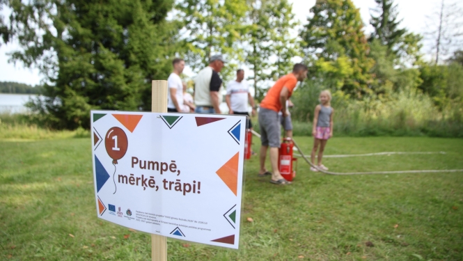 Zālienā novietots plakāts, uz kura uzraksts "Pumpē, mērķē, trāpi!". Fonā rindā sastājušies cilvēki, kur pirmais pumpē ūdeni. Tālāk fonā koki un krūmi.