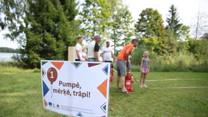 Plakāts, uz kura uzraksts "Pumpē, mērķē, trāpi!". Fonā rindā sastājušies cilvēki un koki.