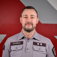 Vīrietis, kas tērpies pelēkā formastērpā, skatās fotogrāfa virzienā.