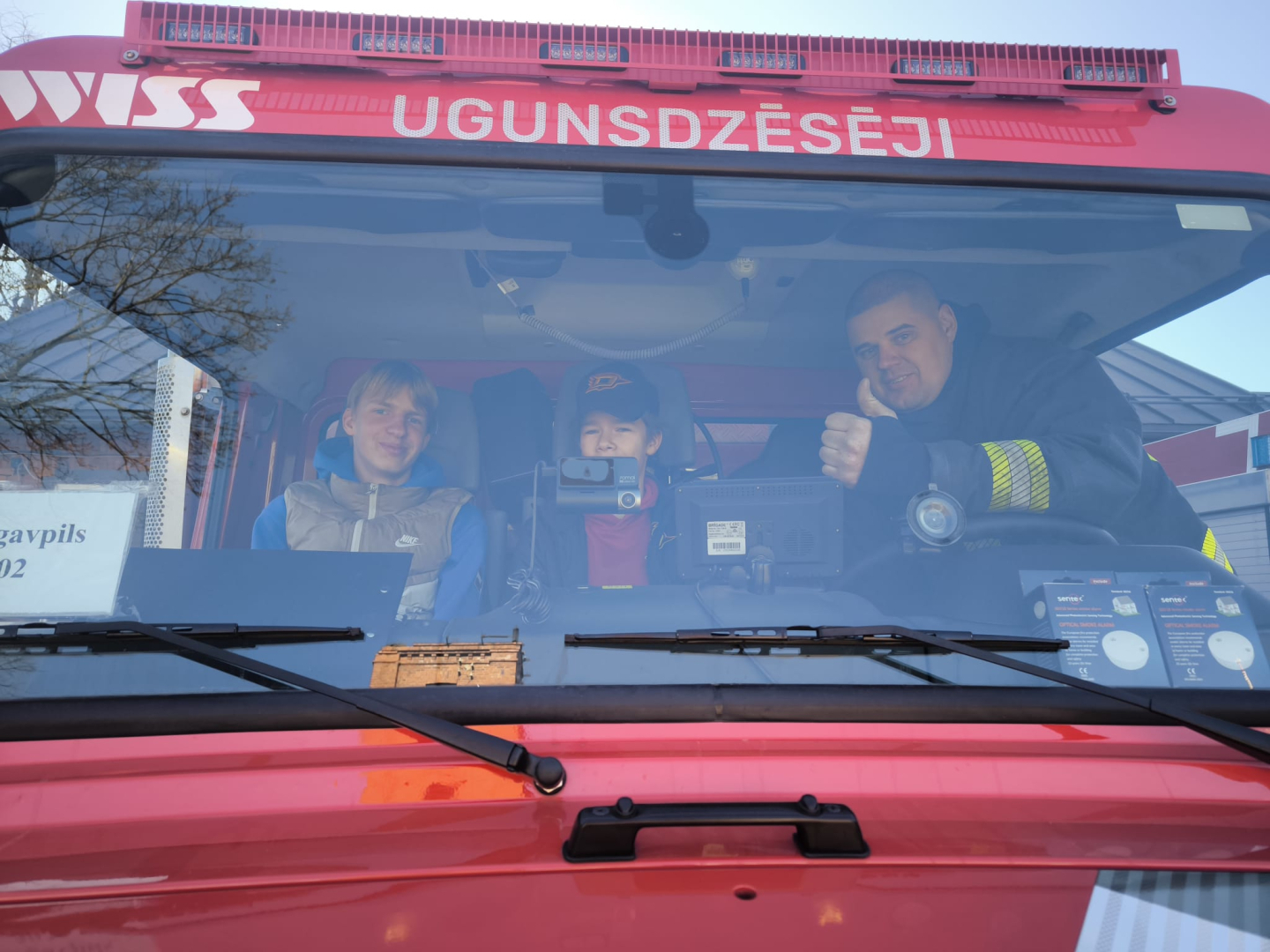 Ugunsdzēsēju glābēju autocisternas priekšējos sēdekļos sēž divi jaunieši un viens ugunsdzēsējs glābējs. Visi skatās fotogrāfa virzienā. Ugunsdzēsējs glābējs ar augšup paceltu īkšķi rāda, ka viss ir labi.