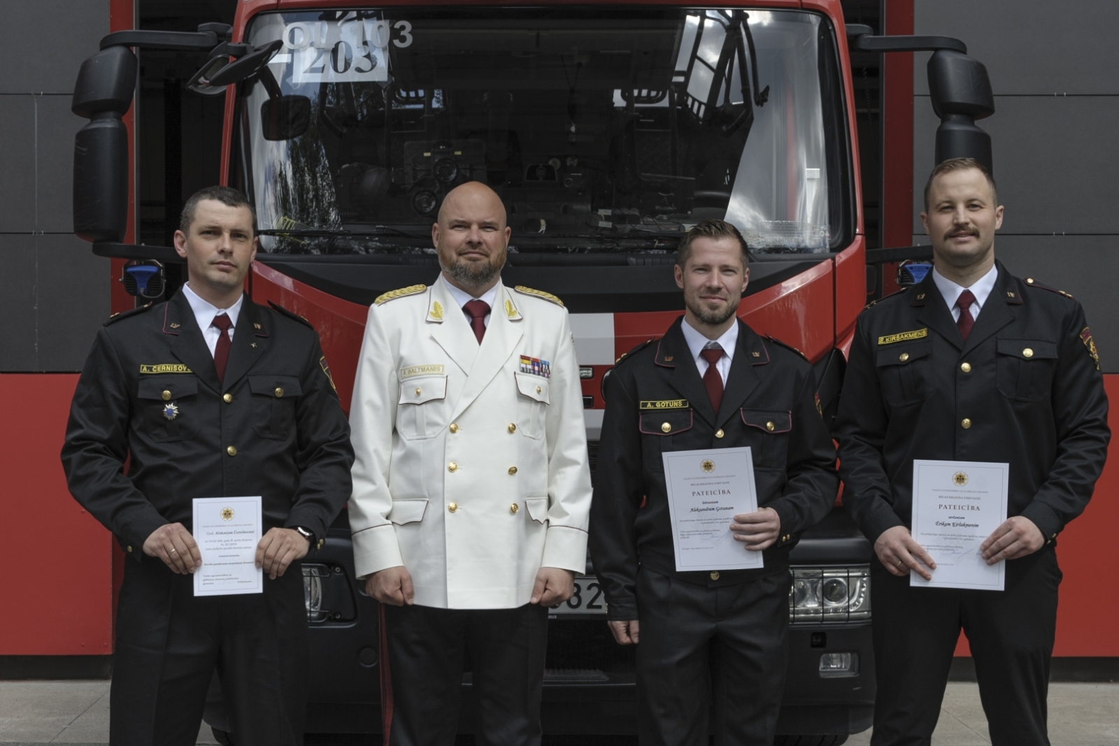 VUGD priekšnieks Mārtiņš Baltmanis un trīs VUGD nodarbinātie, kuriem rokās atzinības raksti. Aiz muguras dienesta specializētais transportlīdzeklis.