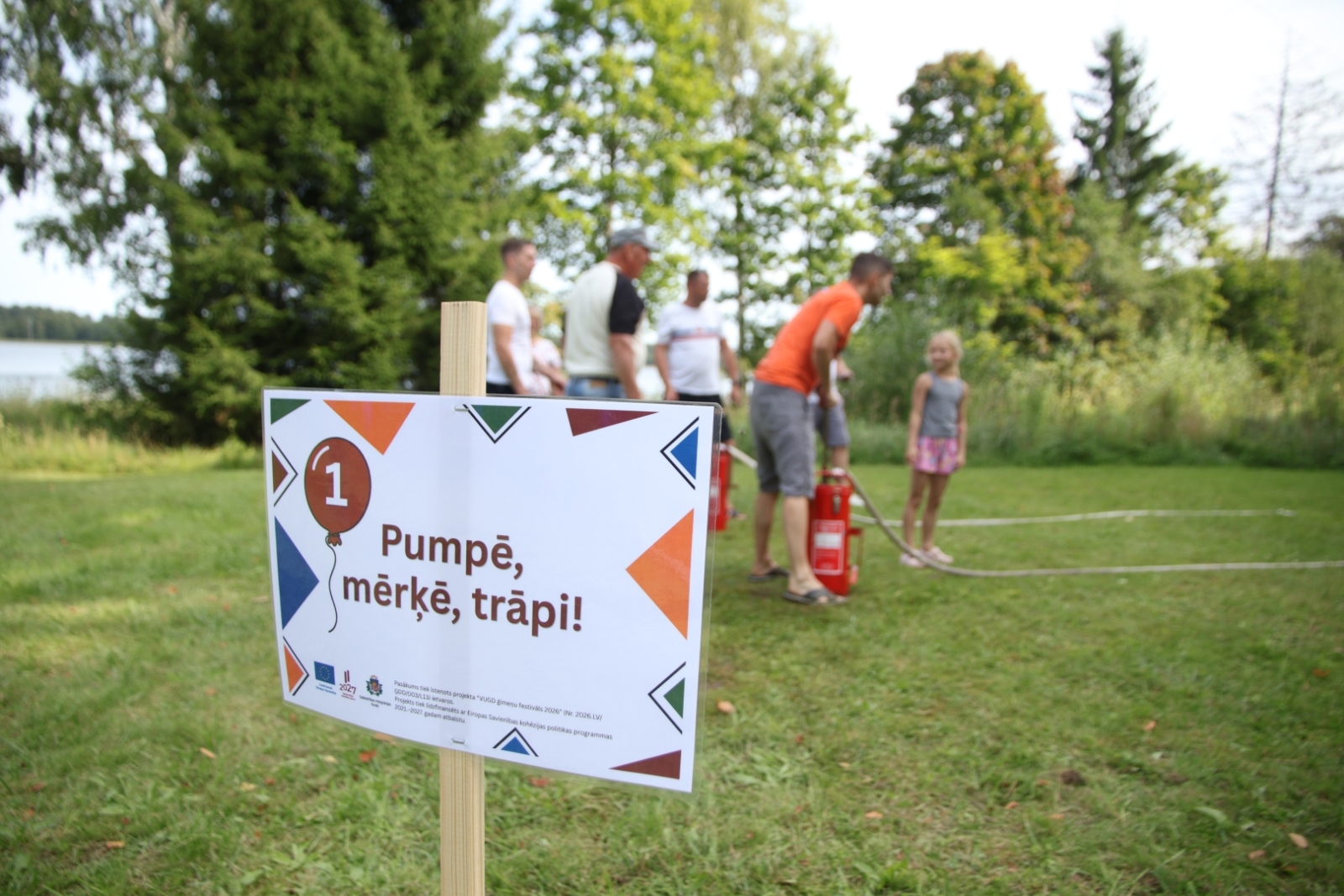 Plakāts, uz kura uzraksts "Pumpē, mērķē, trāpi!". Fonā rindā sastājušies cilvēki un koki.