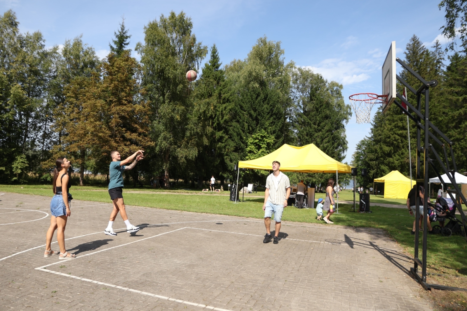 Vīrietis met basketbola grozā bumbu. Kreisajā pusē stāv sieviete, labajā vīrietis. Fonā dzeltena telts un koki.