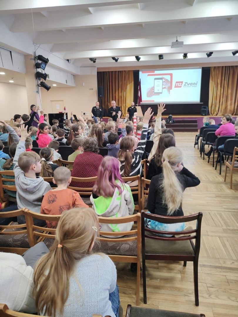 Zālē sēž daudz jaunieši un skatās uz zāles skatuves izvietoto prezentāciju 112. Daži bērni ir pacēluši rokas