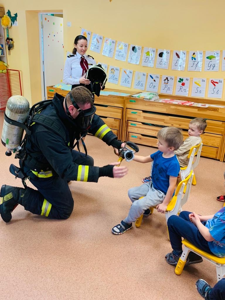 Ugunsdzēsējs glābējs notupies uz ceļa rāda bērnam, kurš sēž uz ķeblīša, vienu no ugunsdzēsēju glābēju instrumentiem