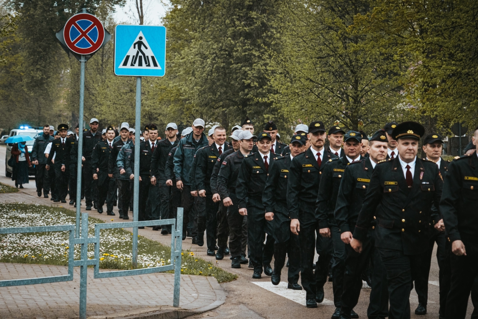 VUGD nodarbinātie kolonnās iet pa ielu. Kreisajā pusē divas ceļa zīmes, kas apzīmē gājēju pāreju un "apstāties, stavēt aizliegts"