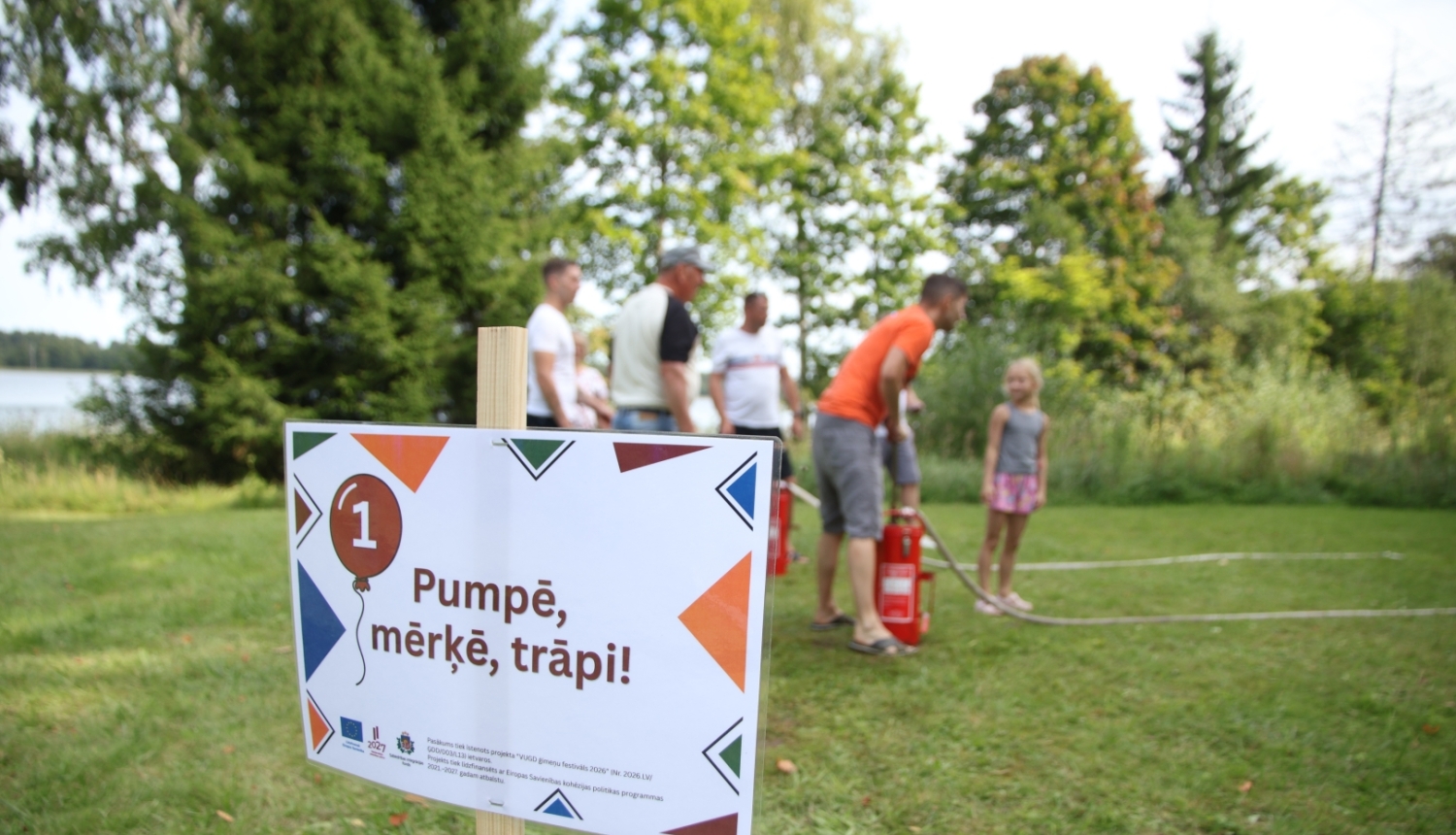 Plakāts, uz kura uzraksts "Pumpē, mērķē, trāpi!". Fonā rindā sastājušies cilvēki un koki.