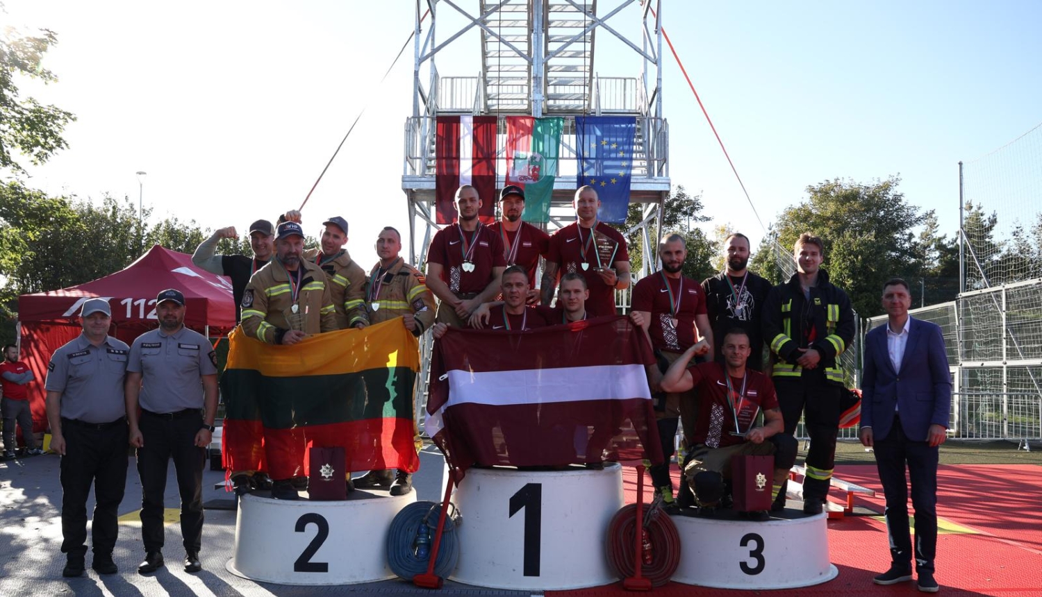 Komandu sacensību dalībnieki pozē uz goda pjedestāla. Vidējā grupa tur rokās Latvijas karogu, grupa pa labi tur Lietuvas karogu. 