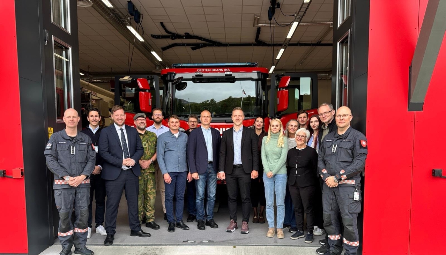 Atvērtas lielas un sarkanas ugunsdzēsības depo garāžas durvis, pie tām stāv vizītes dalībnieki un skatās fotogrāfa virzienā. Fonā redzama ugunsdzēsēju glābēju autocisternas augšējā daļa - priekšējais stikls, spoguļi u.tml.