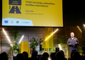 Uz skatuves stāv vīrietis formastērpā. Pie sienas piestiprināts liels ekrāns. Uz ekrāna redzams prezentācijas slaids dzeltenā krāsā.