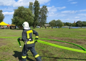 Pa zaļu zālienu iet ugunsdzēsējs glābējs, kurš rokās tur spilgti zaļas krāsas ūdens šļūteni. Uz zemes zālienā novietotas vēl citas trīs spilgti zaļas ūdens šļūtenes. Fonā redzami zaļi koki, dzeltena telts un zilas debesis