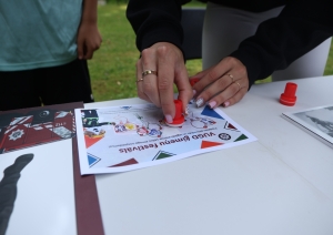 Redzamas sievietes rokas, kas uz lapas, kura atrodas uz galda, uzspiež zīmogu. Uz lapas uzraksts "VUGD ģimeņu festivāls". Blakus sievietei kreisajā pusē redzams, ka stāv vēl kāda persona.