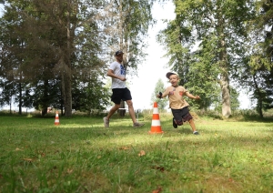 Zālienā nolikti divi konusi, ap tiem skrien vīrietis un bērns. Fonā koki.