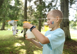 Bērns ar ūdens smidzināmo pudeli smidzina ūdeni plastmasas krūzītē. Kūzīte ievērta striķī, kas nostiprināts starp kokiem. Fonā citi koki. 