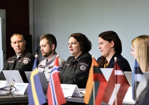 Pie galda sēž pieci darba grupas dalībnieki. Priekšplānā nolikti karogi.