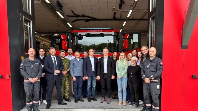 Atvērtas lielas un sarkanas ugunsdzēsības depo garāžas durvis, pie tām stāv vizītes dalībnieki un skatās fotogrāfa virzienā. Fonā redzama ugunsdzēsēju glābēju autocisternas augšējā daļa - priekšējais stikls, spoguļi u.tml.