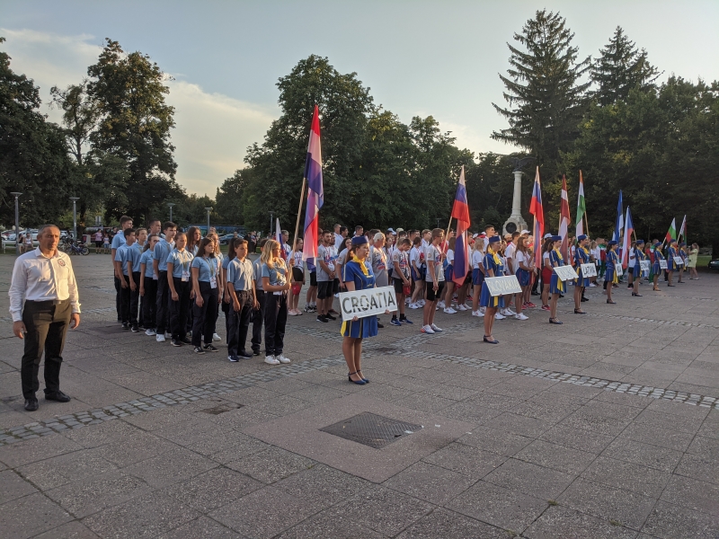 Uz laukuma stāv sacensību dalībnieku komandas. To priekšpusē nostājušās sievietes, tērpušās zili dzeltenos tērpos, kuras rokās tur baltus plakātus uz kuriem melniem burtiem rakstīts konkrētās komandas pārstāvētās valsts nosaukums.