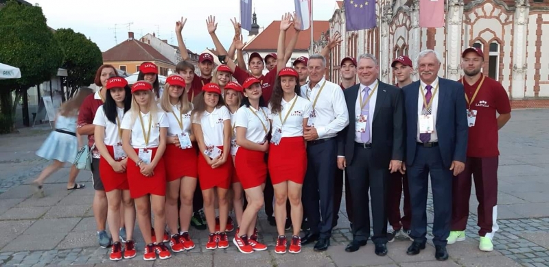Latvijas komandas un viņu pavadoņu kopbilde. Visi skatās fotogrāfa virzienē. Aizmugurē stāvošie jaunieši pacēluši rokas uz augšu.