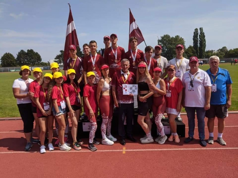 Uz sporta stadiona sarkanā skrejceļa nostājušies sacensību dalībnieki un viņu pavadoņi. Visi skatās fotogrāfa virzienā. Viņiem tuvumā novietoti divi Latvijas Republikas saraknbaltsarkanie karogi.
