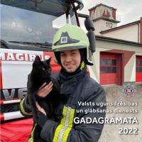 Ugunsdzēsējs glābējs tērpies savā aizsargtērpā stāv pie ugunsdzēsēju glābēju mašīnas un tur rokās melnu kaķi. Fonā redzams ugunsdzēsēju glābēju depo