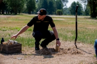 Ugunsdzēsējs glābējs pietupies pie zemes. Ar vienu roku liek smiltīs malkas pagali, bet ar otru roku ņem jaunu malkas pagali no groza. Blakus ugunsdzēsējam glābējam redzama smiltīs iedurta lāpsta.