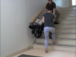 Divi vīrieši palīdz pārvietoties personai ratiņkrēslā augšup pa kāpnēm