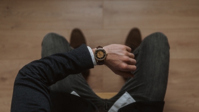 Cilvēks uzvalkā sēž un skatās uz rokas pulksteni. Fonā redzama brūna grīda
