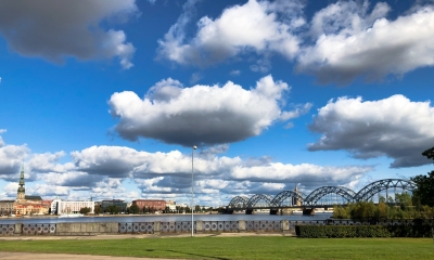 Zilas debesis ar baltiem mākoņiem, zaļa zāle. Tālumā pilsēta - Rīgas dzelzceļa tilts pār Daugavu, Vecrīga