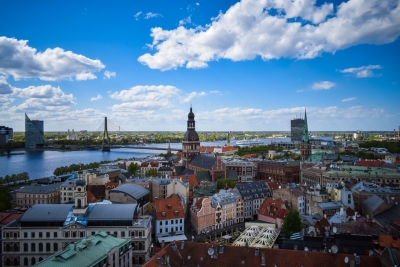 Zilas debesis ar baltiem mākoņiem. Zem tām Rīgas mājas un torņu smailes. Tilts pār Daugavu.