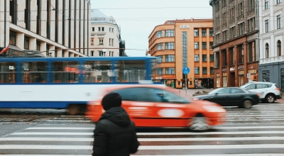 Cilvēks tērpies melnās drēbēs stāv pie gājēju pārejas. Garām brauc sarkana mašīna, zili baltais trolejbuss. Tālumā redzamas dažādas mājas