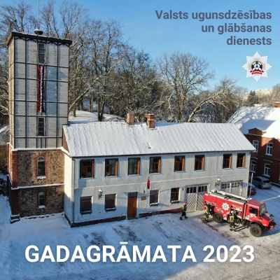 Baltas krāsas ugunsdzēsības depo. Tam kreisajā malā augsts tornis. Pie depo novietota sarkana VUGD autocisterna, tai blakus stāv ugunsdzēsēji glābēji. Apakšā teksts Gadagrāmata 2023