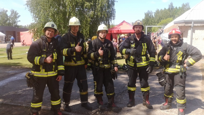 Pieci ugunsdzēsēji glābēji stāv ietērpušies savos formas tērpos un skatās fotogrāfa virzienā