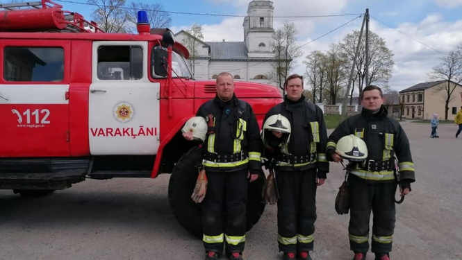 Trīs ugunsdzēsēji glābēji stāv pie vienas ugunsdzēsēju glābēju mašīnas un ietur Klusuma brīdi