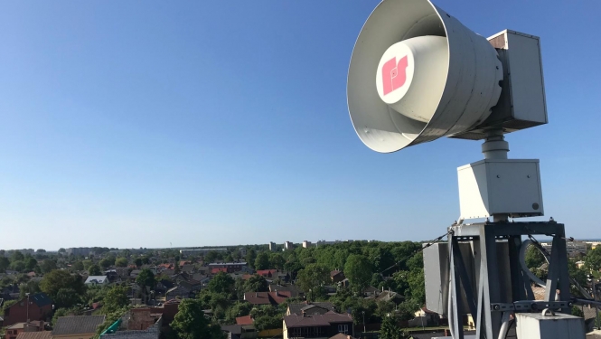 Uz jumta izvietota balta trauksmes sirēna. Tālumā redzamas zilas debesis, māju jumti un zaļas koku galotnes.