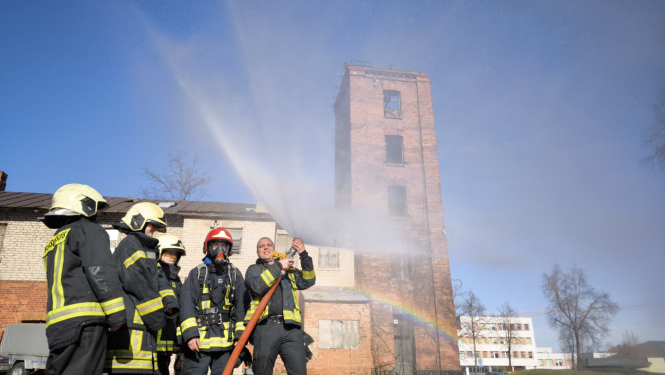 Jaunieši, kas tērpušies ugunsdzēsēju glābēju aizsargtērpos stāv pagalma vidū. Ugunsdzēsējs glābējs tur rokās uz augšu paceltu ugunsdzēsības šļūteni, no kuras uz visām pusēm smidzinās ūdens, kas saules staros veido varavīksni.