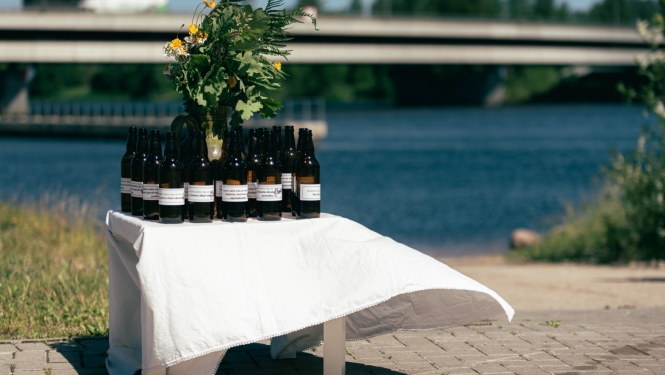 Uz maza galdiņa, kas apklāts ar baltu galdautu, novietotas tumši brūnas atvērtas alus pudeles un jāņuzāļu pušķis. Alus pudelēm uzlīmētas etiķetes ar aicinājumu aizdomāties par to, lai svētkus pavadītu droši