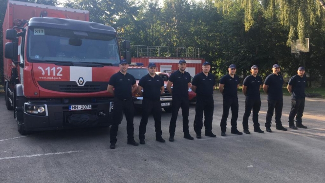 Astoņi vīrieši, tērpušies formastērpos, stāv vienā taisnā līnijā viens otram blakus un skatās fotogrāfa virzienā. Aiz viņiem novietoti VUGD sarkanbaltie transportlīdzekļi