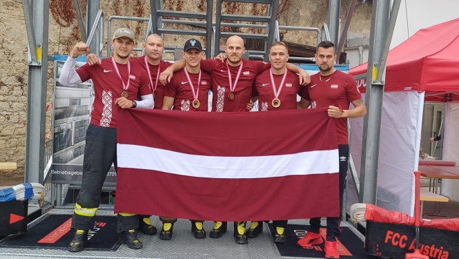 Seši vīrieši, tērpušies sarkanos kreklos un pelēkās biksēs, tur rokās Latvijas karogu un skatās fotogrāfa virzienā. Vīriešiem kaklā uzkārtas bronzas medaļas