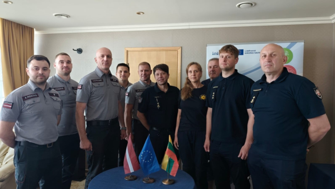 Desmit Latvijas un Lietuvas operatīvo dienestu nodarbinātie stāv ap mazu galdiņu uz kura novietoti trīs karogi - Latvijas, Igaunijas un Lietuvas