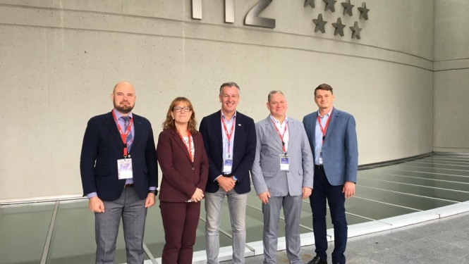 Piecu cilvēku - vienas sievietes un četru vīriešu - kopbilde. Visi stāv un skatās fotogrāfa virzienā. Fonā redzama siena uz kuras ir redzams tālruņa numurs 112 un septiņas zvaigznes.