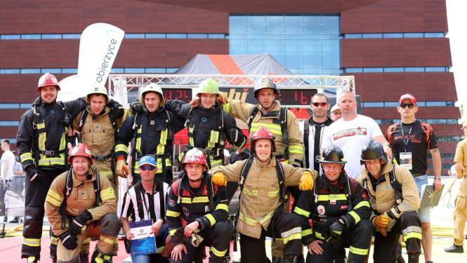 Četrpadsmit vīriešu kopbilde. Desmit vīrieši tērpušies ugunsdzēsēju glābēju apģērbā - zilās formās ar dzeltenas krāsas atstarotājiem un bēšās formās ar dzeltenas krāsas atstarotājiem. Visiem ugunsdzēsējiem glābējiem galvā uzvilktas ķiveres - baltas un sarkanas.