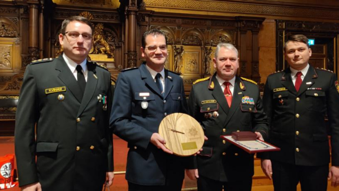 Četri vīrieši tērpušies formastērpos stāv un skatās fotogrāfa virzienā. Viens vīrietis rokās tur izgrebtu koka apli