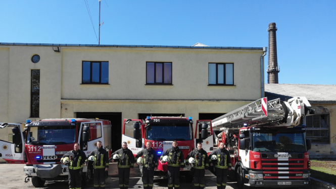 Septiņi ugunsdzēsēji glābēji stāv, turot rokās savas ķiveres. Viņiem fonā redzamas trīs dažādas ugunsdzēsēju glābēju autotehnikas