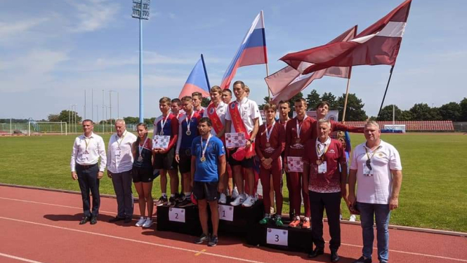 Uz stadiona sarkanīgā skrejceļa novietoti pjedestāli. Uz tiem uzkāpuši sacensību dalībnieki un uzvarētāji. Tiem aiz muguras novietoti trīs valstu karogi.