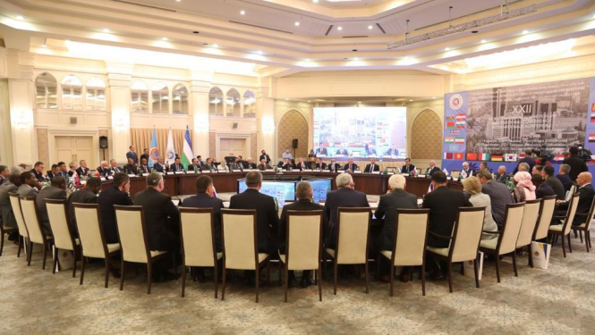 Lielā baltā zālē ar izgaismotiem griestiem novietots liels apaļš galds. Ap to sasēdušies sanāksmes dalībnieki.