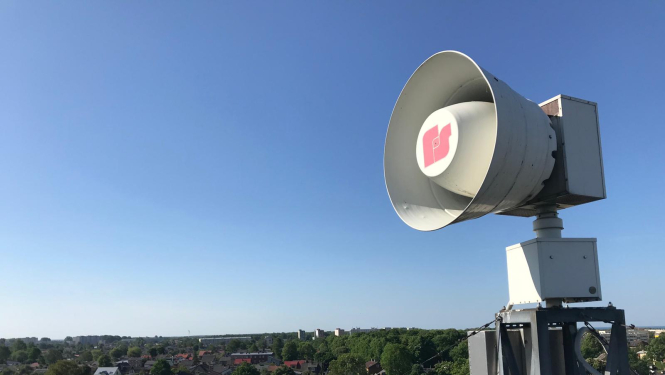 Uz jumta izvietota balta trauksmes sirēna. Tālumā redzamas zilas debesis, māju jumti un zaļas koku galotnes.