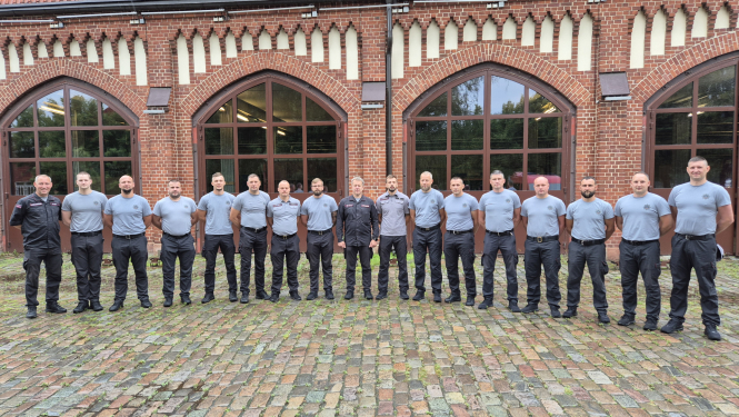 Septiņpadsmit VUGD pelēkajos formastērpos ģērbušies vīrieši stāv uz bruģa un skatās fotogrāfa virzienā. Aiz viņiem atrodas sarkana ķieģeļu māja ar lieliem stiklotiem arkveida logiem.