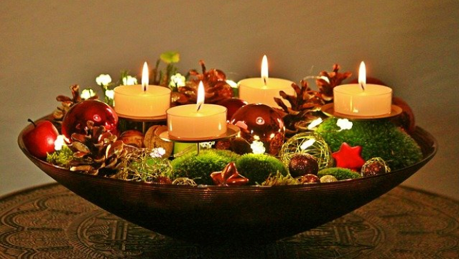 Uz brūna galda novietota lēzena stikla bļoda. Tajā ievietoti dažādi dekoratīvi elementi - čiekuri, plastmasas bumbiņas, zvaigznītes, ābols u.tml., kā arī ievietotas četras dzeltenas svecītes. Visas ievietotās sveces ir aizdegtas.